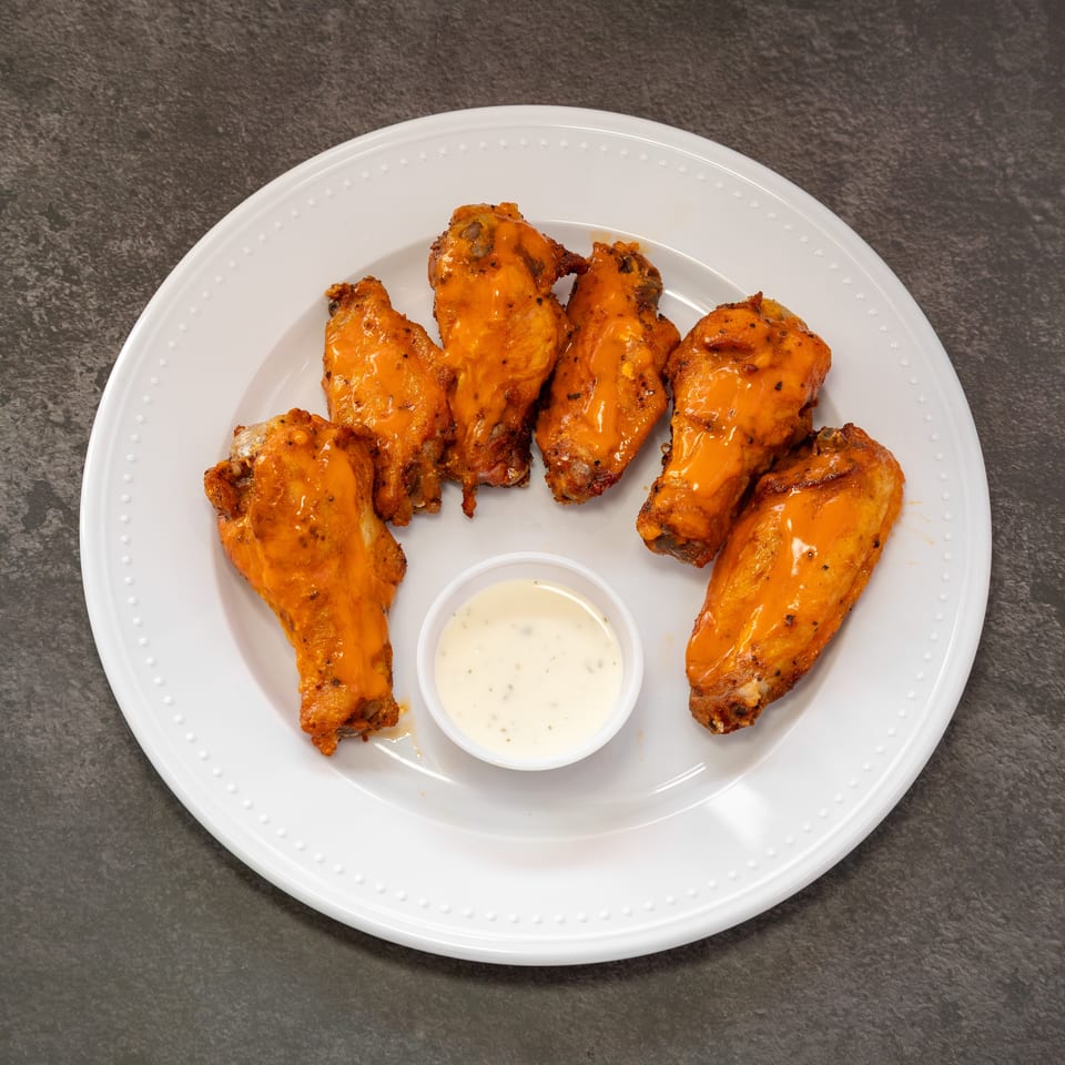 Six buffalo chicken wings with a side of ranch dressing on a white plate.