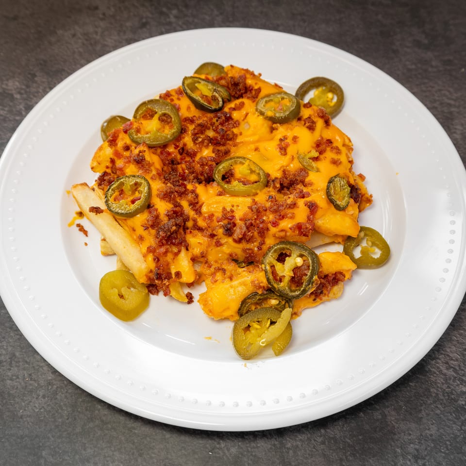 Loaded cheese fries topped with crispy bacon bits and sliced jalapeños on a white plate.