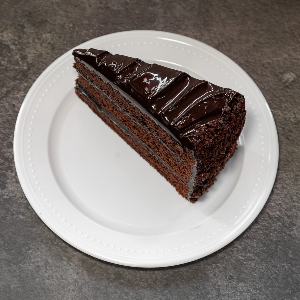 Slice of rich chocolate cake with glossy ganache frosting on a white plate.