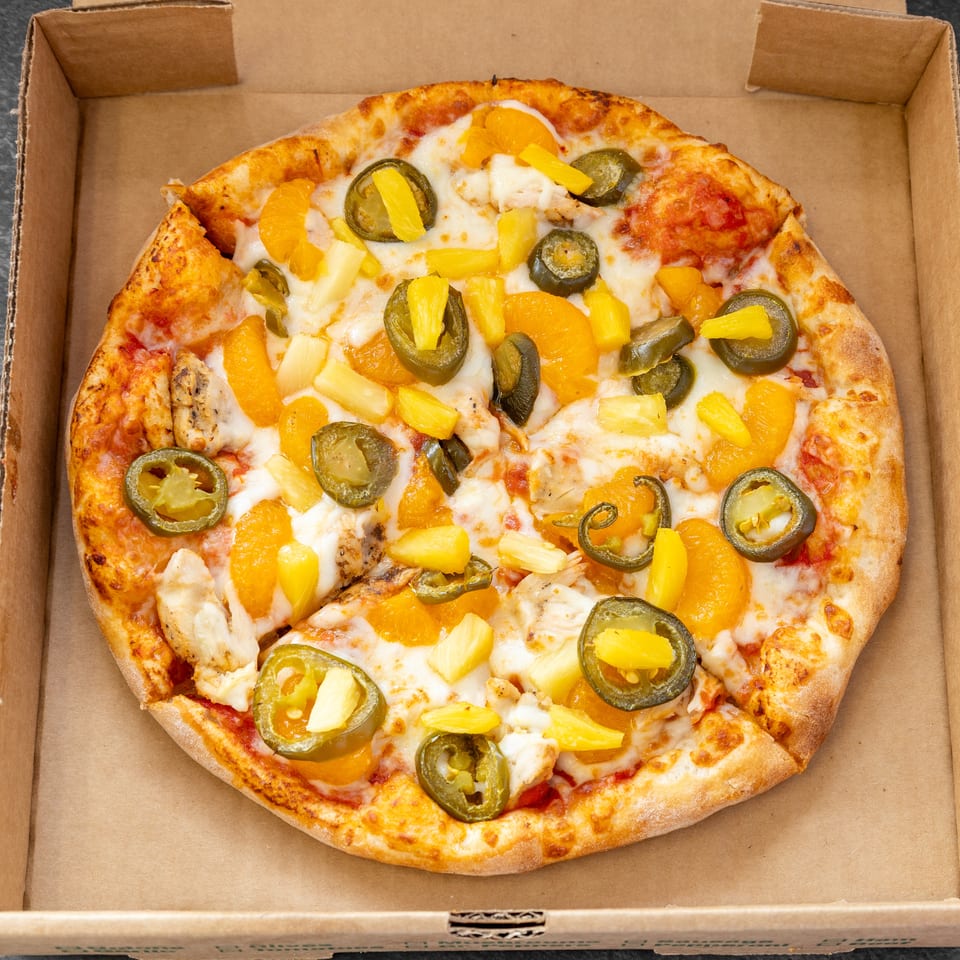 A whole pizza with chicken, pineapple, mandarin oranges, and jalapeños in a cardboard box.