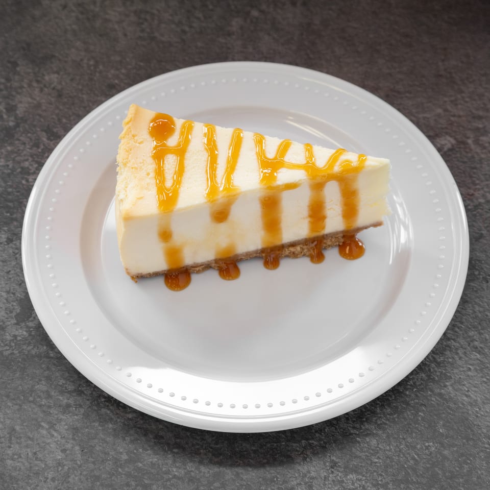 A slice of cheesecake with a graham cracker crust, drizzled with caramel sauce on a white plate.
